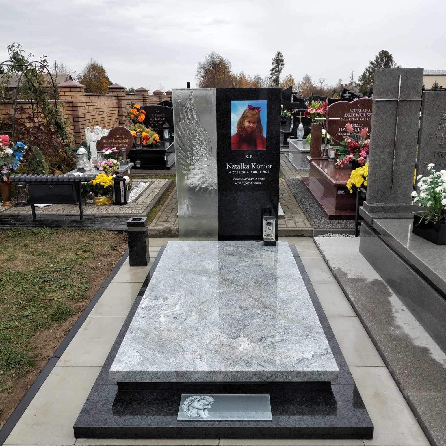 Granitowy nagrobek z portretem dziecka i motywem skrzydła, otoczony innymi nagrobkami na cmentarzu.