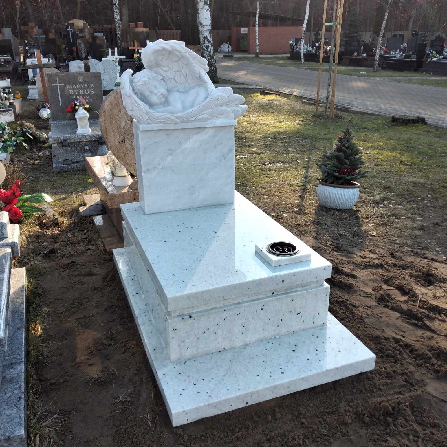 Jasny, trzyczęściowy pomnik z rzeźbą śpiącego anioła na górze, otoczony innymi nagrobkami na cmentarzu, z małą choinką w doniczce w tle.