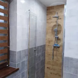 Wykończona łazienka z kabiną prysznicową typu walk-in, ściany wyłożone płytkami w odcieniach szarości i bieli, drewnopodobna okładzina w strefie prysznica, drewniane żaluzje w oknie.