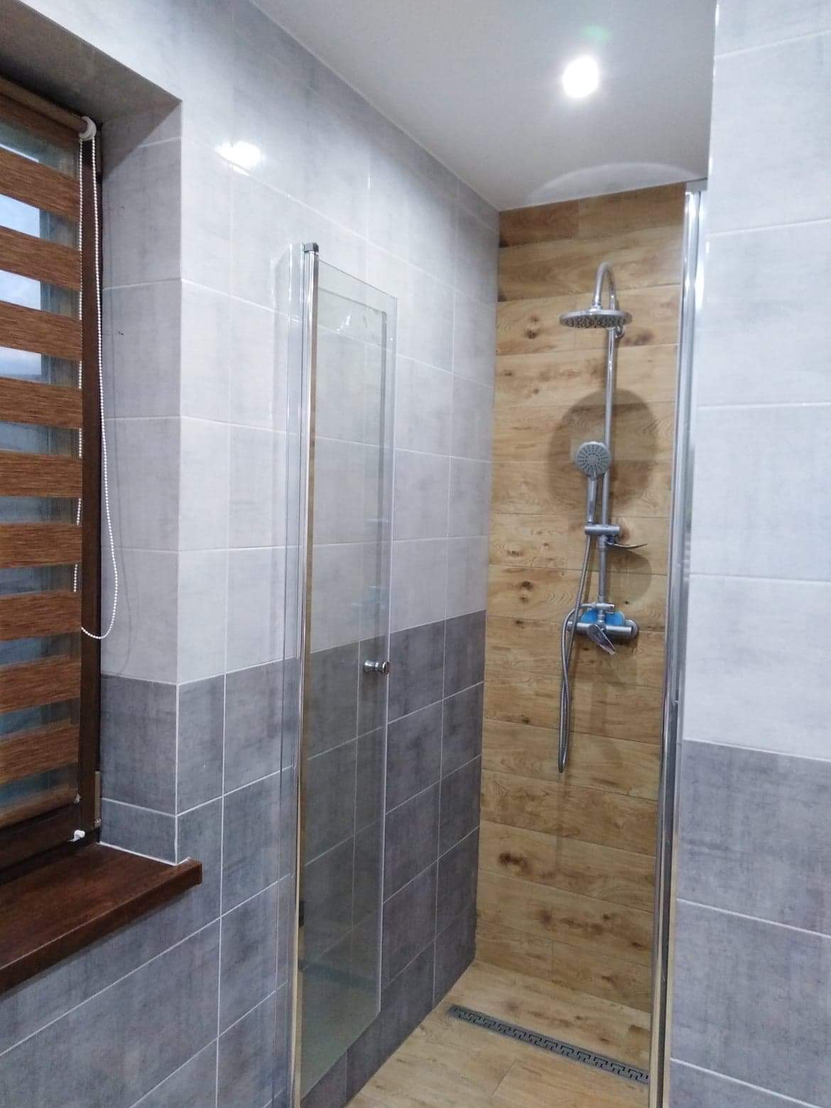 Wykończona łazienka z kabiną prysznicową typu walk-in, ściany wyłożone płytkami w odcieniach szarości i bieli, drewnopodobna okładzina w strefie prysznica, drewniane żaluzje w oknie.