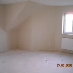 Poddasze w Krakowie po gładziowaniu ścian i sufitu, przygotowane do dalszych prac wykończeniowych. Widoczne gniazdka elektryczne i grzejnik. Na podłodze resztki materiałów budowlanych.