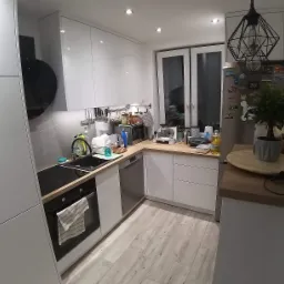 Nowoczesna kuchnia z białymi frontami, drewnianymi blatami i czarnymi akcentami, takimi jak okap i geometryczna lampa wisząca, widok na okno i liczne akcesoria kuchenne na blacie.