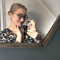 Odbicie w lustrze kobiety w okularach, trzymającej telefon w obudowie z wizerunkiem Królewny Śnieżki, dotykającej szyi dłonią.