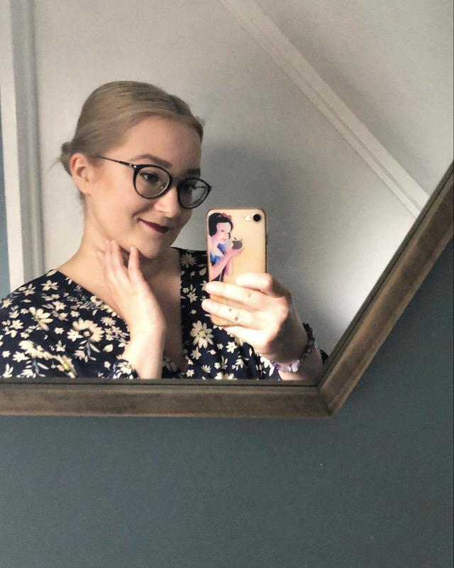 Odbicie w lustrze kobiety w okularach, trzymającej telefon w obudowie z wizerunkiem Królewny Śnieżki, dotykającej szyi dłonią.