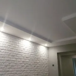 Ściana wykończona białą cegłą dekoracyjną, oświetlona punktowymi lampami w suficie podwieszanym z gładzią, widok na fragment korytarza z szarą ścianą.