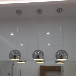 Biała, błyszcząca łazienka z trzema okrągłymi lampami chromowanymi wiszącymi nad lustrem, odbijającymi światło.