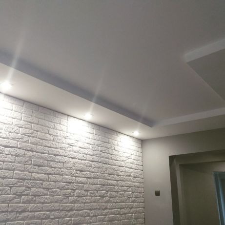 Ściana wykończona białą cegłą dekoracyjną, oświetlona punktowymi lampami w suficie podwieszanym z gładzią, widok na fragment korytarza z szarą ścianą.