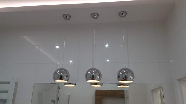 Biała, błyszcząca łazienka z trzema okrągłymi lampami chromowanymi wiszącymi nad lustrem, odbijającymi światło.