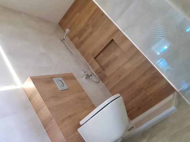 Nowoczesna łazienka z drewnopodobnymi płytkami na ścianie, podświetlona listwa LED, wisząca toaleta i wnęka w ścianie wykończona tym samym materiałem.