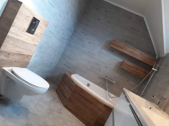 Nowoczesna łazienka z drewnopodobnymi płytkami, wisząca toaleta z chromowanym przyciskiem, wanna obudowana drewnem, szare płytki na podłodze i ścianach, drewniane półki.