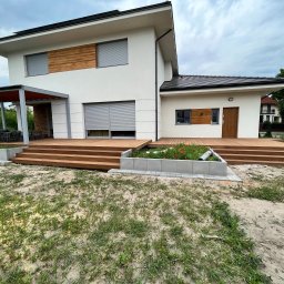 DKS Drewniane Konstrukcje Szkieletowe MARCIN BADOWSKI - Nowoczesny dom z jasną elewacją i rozległym tarasem z drewna egzotycznego, prowadzącym do wejścia, z widoczną pergolą z metalową konstrukcją i drewnianym zadaszeniem, oraz rabatą kwiatową z przodu.