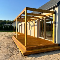 DKS Drewniane Konstrukcje Szkieletowe MARCIN BADOWSKI - Nowoczesny taras z drewna zadaszony drewnianą pergolą, przylegający do jasnego domu z dużymi oknami przesuwnymi, widoczne schody wejściowe i niezagospodarowany teren wokół.