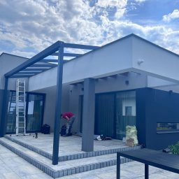 DKS Drewniane Konstrukcje Szkieletowe MARCIN BADOWSKI - Montaż metalowej pergoli przy nowoczesnym domu z szarymi elementami, widoczny pracownik podczas pracy na tarasie z szarej kostki brukowej, drabina oparta o ścianę.