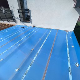 DKS Drewniane Konstrukcje Szkieletowe MARCIN BADOWSKI - Montaż przezroczystego, niebieskiego zadaszenia tarasu z poliwęglanu komorowego, zabezpieczonego taśmą z napisem FACE U.V., widoczne drewniane legary konstrukcji nośnej oraz narzędzia...