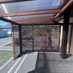 DKS Drewniane Konstrukcje Szkieletowe MARCIN BADOWSKI - Zadaszona, drewniana konstrukcja z lamelami bocznymi, oświetlona sznurem żarówek, stojąca na szarym brukowanym podłożu, widok z zewnątrz w słoneczny dzień.