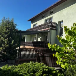 Dwukondygnacyjny dom z balkonem i tarasem z ciemną, nowoczesną pergolą. Widok z ogrodu w słoneczny dzień. Elementy drewniane i roślinność.