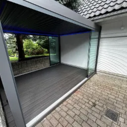 Nowoczesna, szara pergola z oświetleniem LED i szklanymi ścianami, przylegająca do budynku z roletą. Taras z desek kompozytowych.