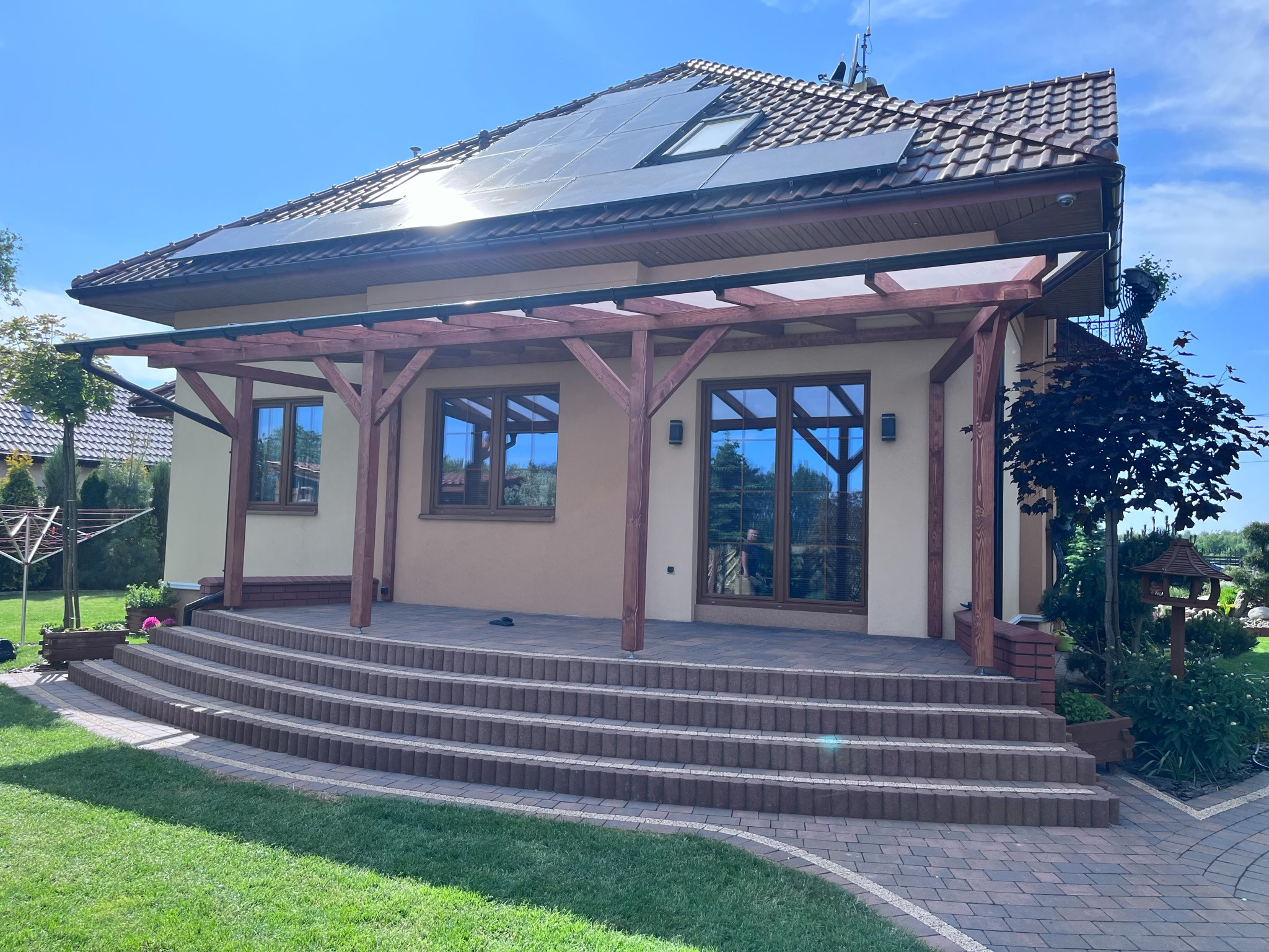 Nowoczesny dom z beżową elewacją, drewnianą pergolą i panelami słonecznymi na dachu, widok na taras z brukowanymi schodami i zadbany trawnik.