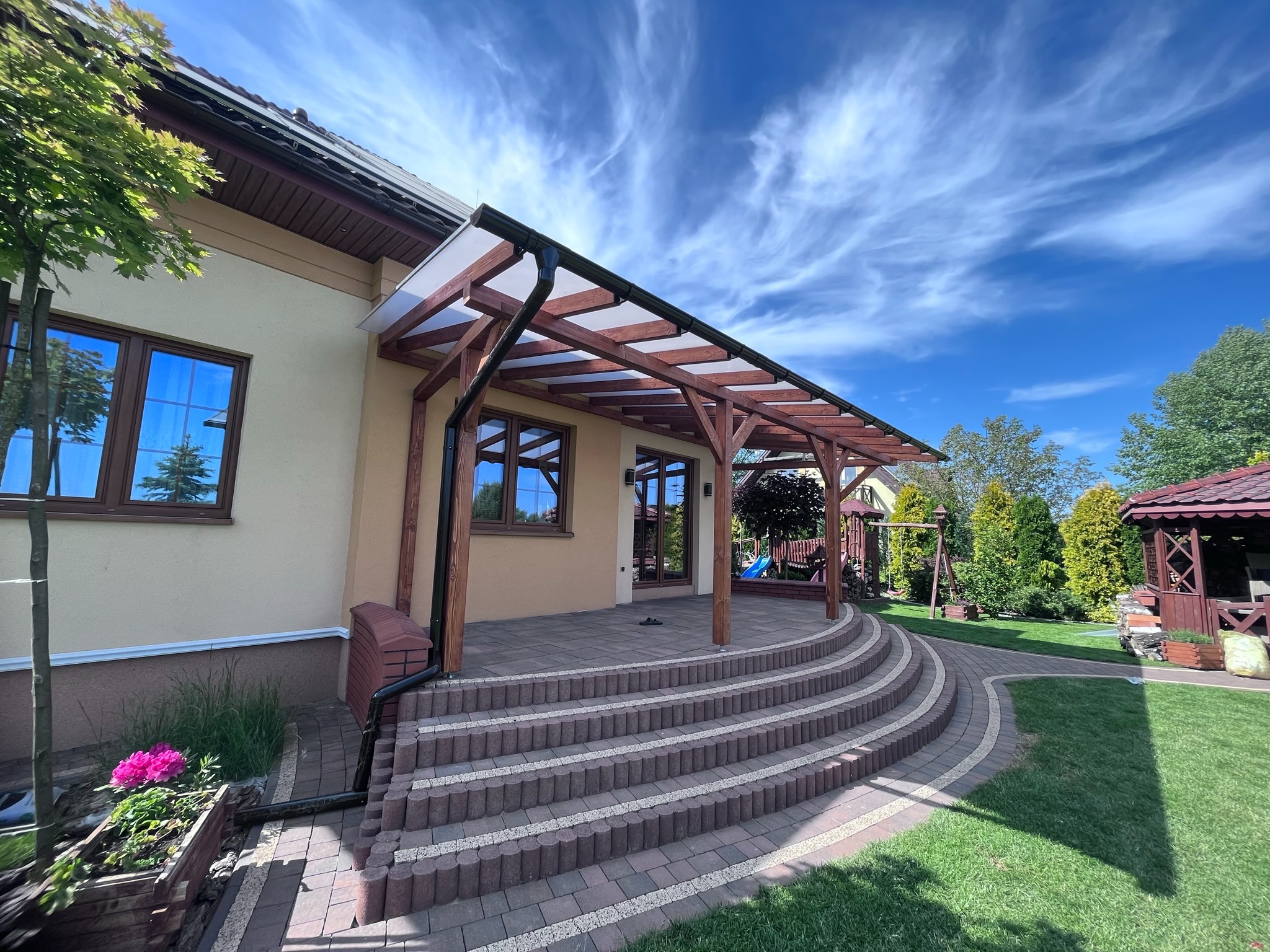 Drewniana pergola z przezroczystym zadaszeniem nad tarasem z kostki brukowej, schody z obrzeży, widok na ogród z altaną i huśtawką w tle, błękitne niebo z chmurami.