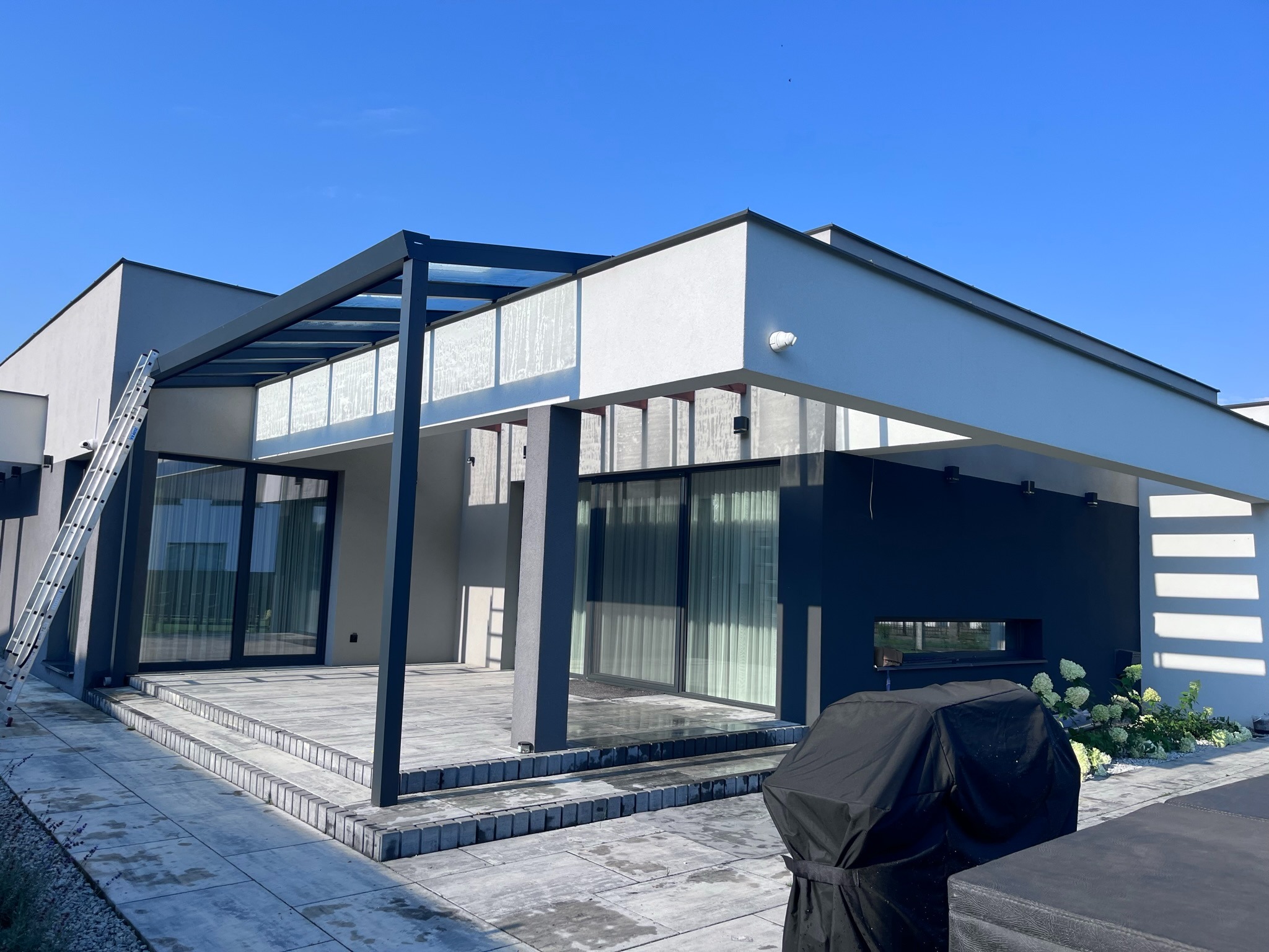 Nowoczesny dom z płaskim dachem i tarasem wyłożonym szarą kostką brukową, z czarną metalową pergolą z przezroczystym zadaszeniem, na tle błękitnego nieba. Przy ścianie domu przystawiona aluminiowa...