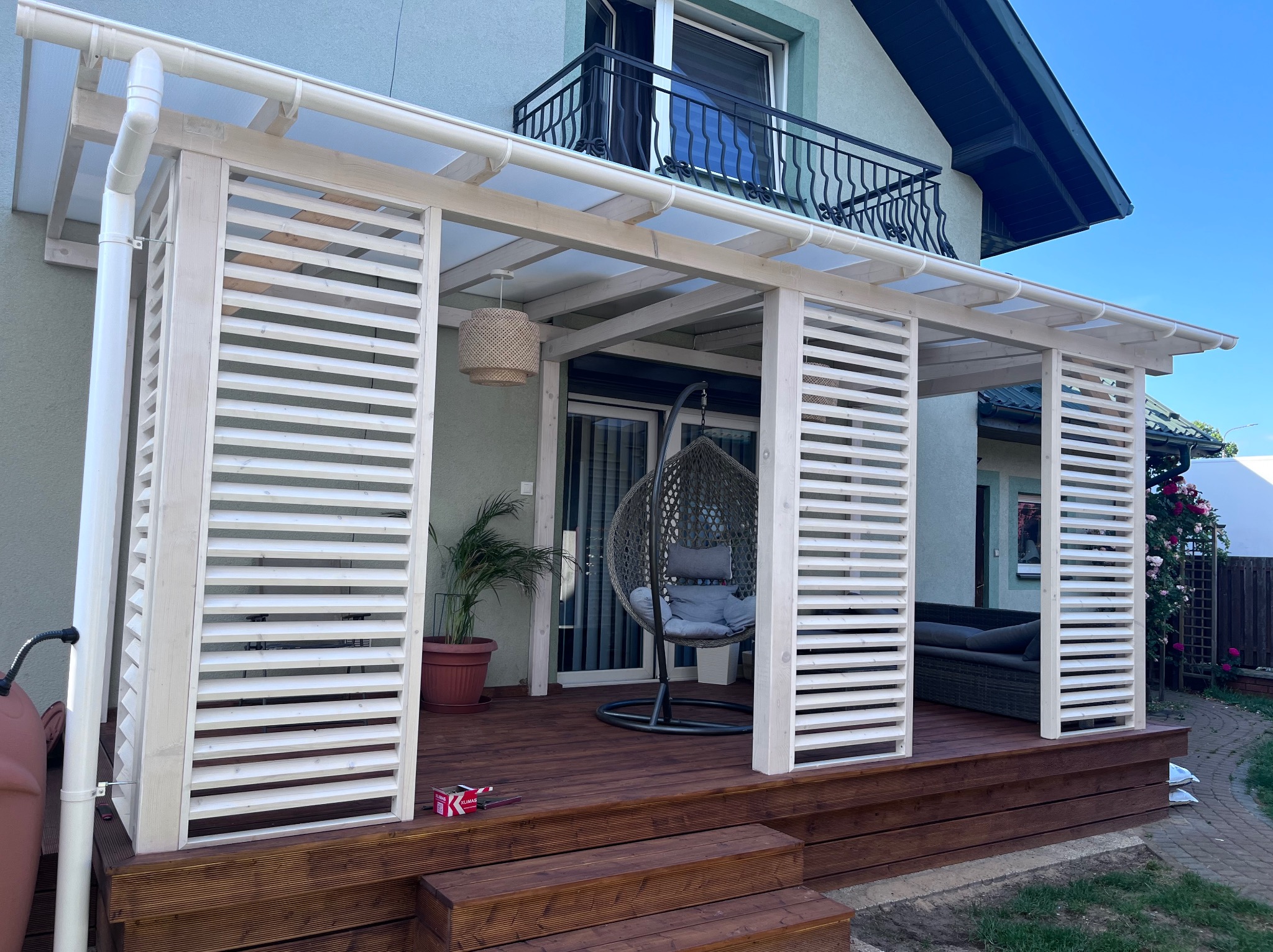 Zadaszony taras z drewnianą konstrukcją, białymi lamelami, wiszącym fotelem i doniczką z rośliną; widoczne schodki wejściowe i fragment domu z balkonem.