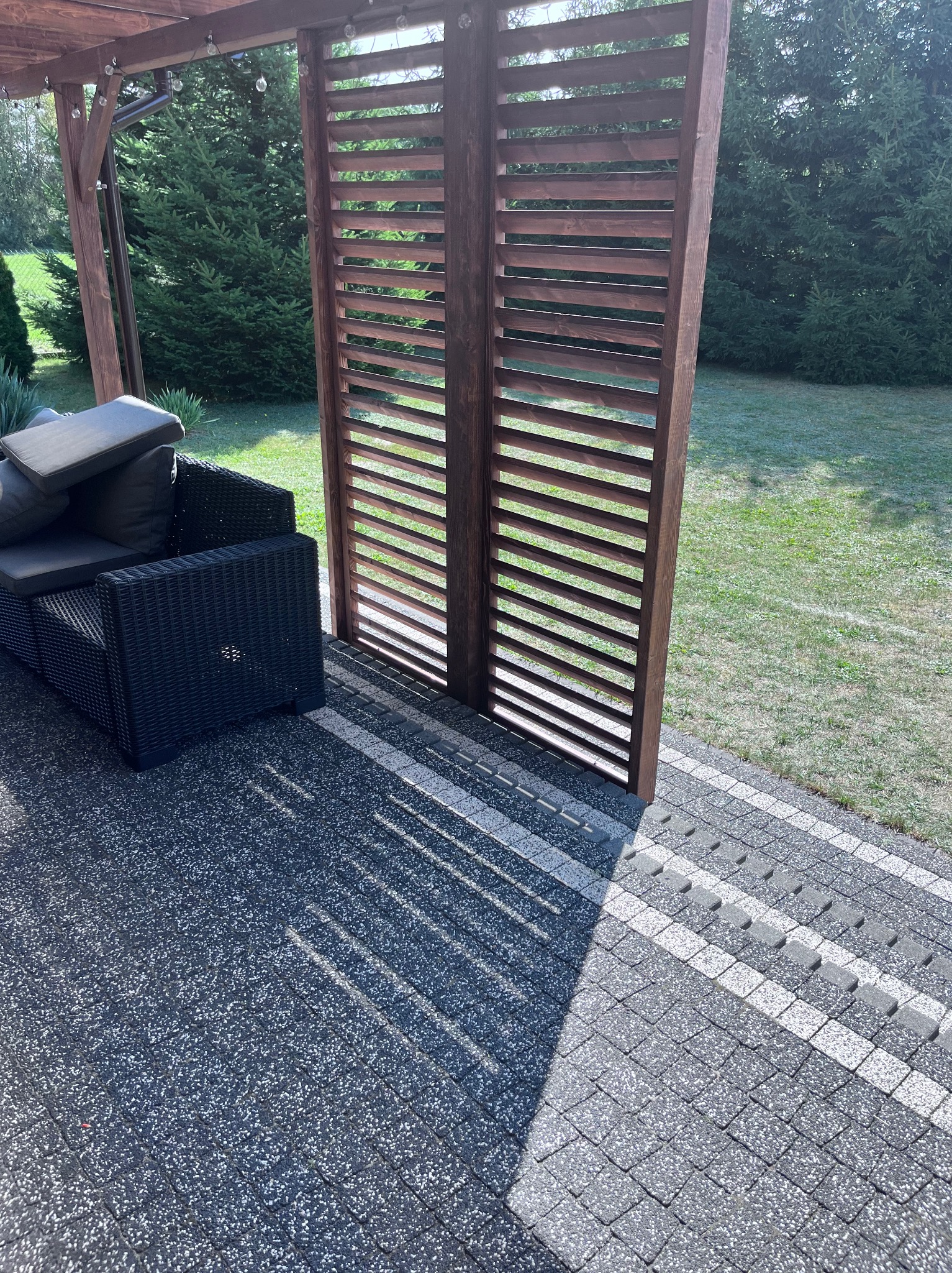 Nowoczesny taras z ciemnoszarą kostką, czarnym rattanowym narożnikiem i drewnianą pergolą ze ścianką z poziomych listew, w tle zielony trawnik i drzewa.