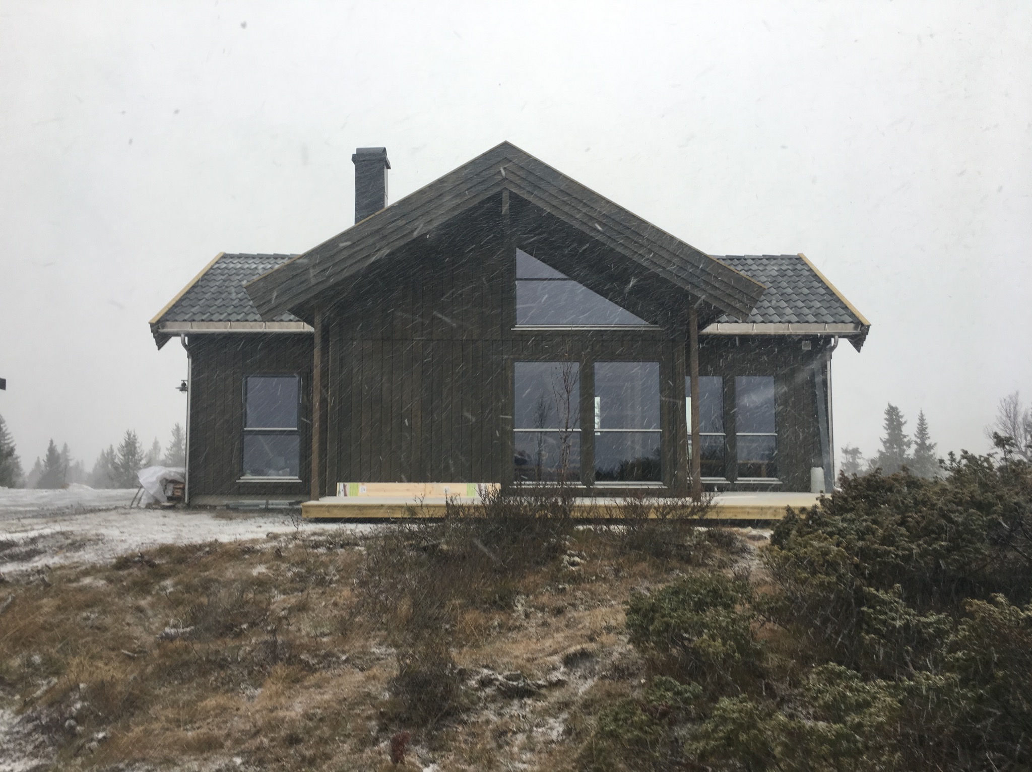 Nowoczesny dom z ciemnego drewna z dużymi oknami i trójkątnym oknem na poddaszu, widoczny w zimowej scenerii podczas opadów śniegu, z fragmentem drewnianego tarasu w budowie.