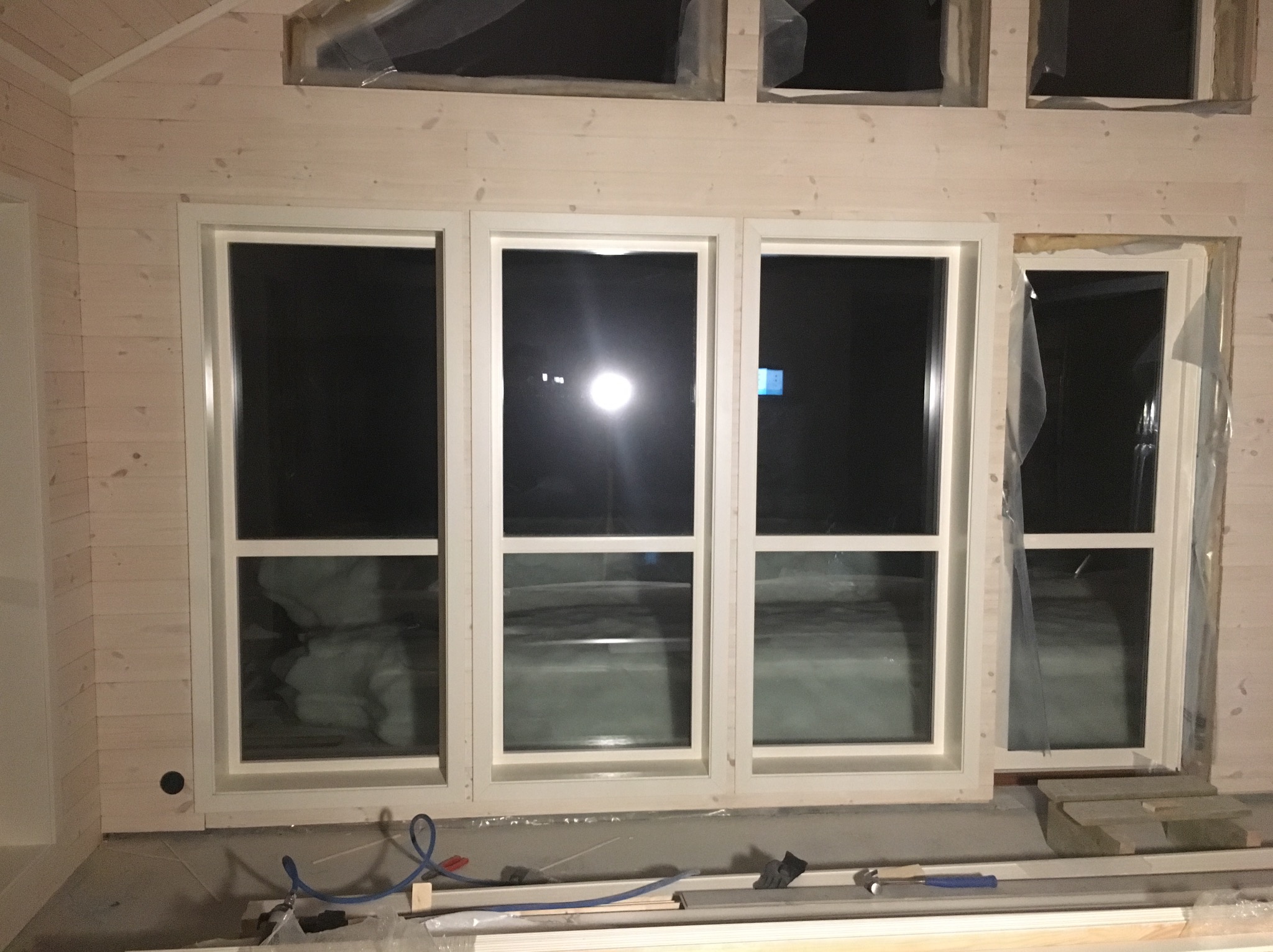 Wnętrze domu w trakcie budowy z drewnianą boazerią i zamontowanymi oknami z białymi ramami, widoczne narzędzia i materiały budowlane.