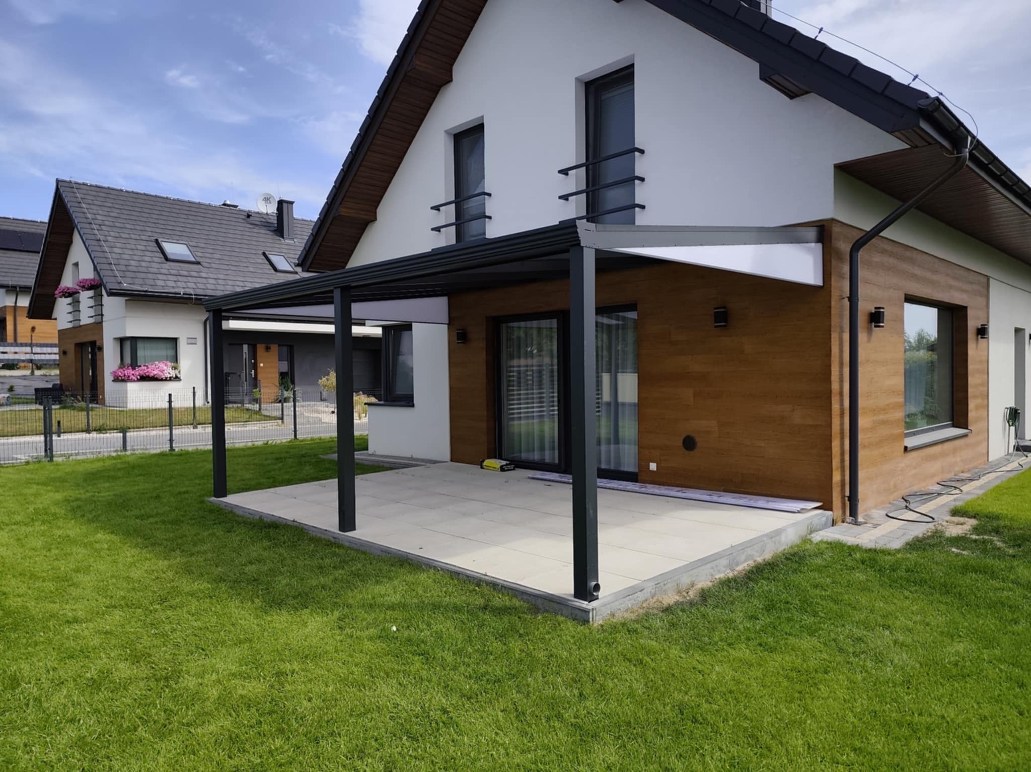 Nowoczesna pergola metalowa z białym zadaszeniem, przylegająca do domu z drewnianą elewacją. Widok na zadbany trawnik i sąsiedni budynek.