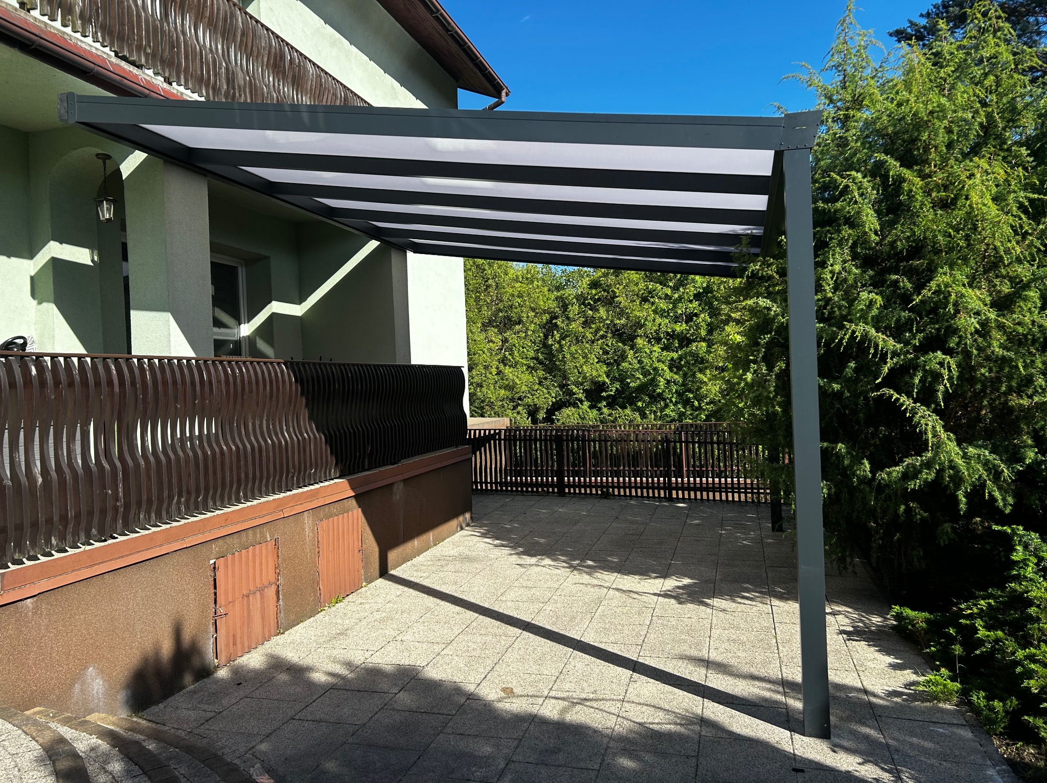 Nowoczesna pergola metalowa z szarym stelażem i białym zadaszeniem, przylegająca do budynku z zieloną elewacją. Widoczny taras z betonowych płyt i zieleń w tle.