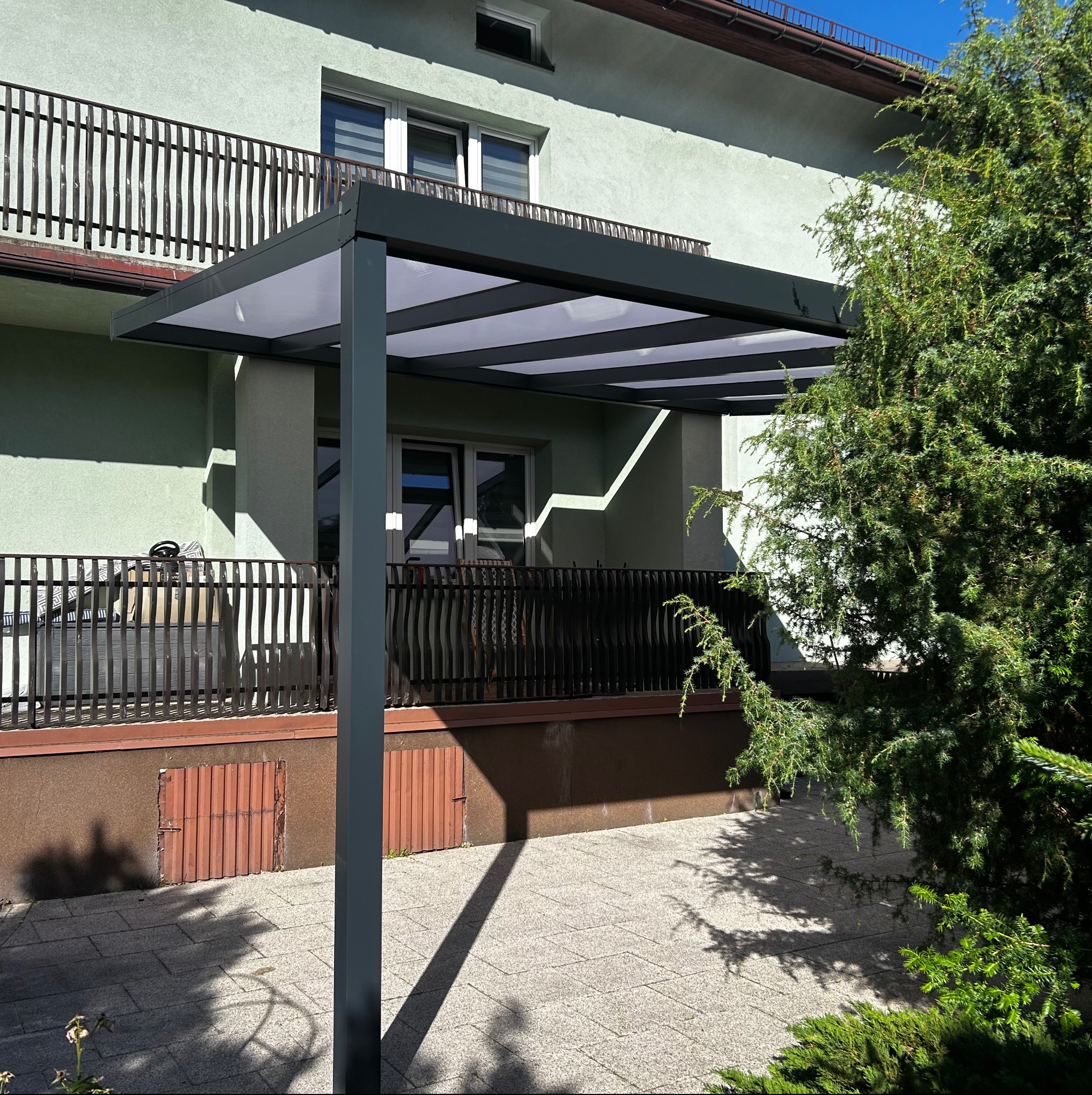 Nowoczesna, szara pergola metalowa z białym zadaszeniem, przylegająca do jasnego budynku. Widok na balkon z ozdobną balustradą i fragment zielonego krzewu.