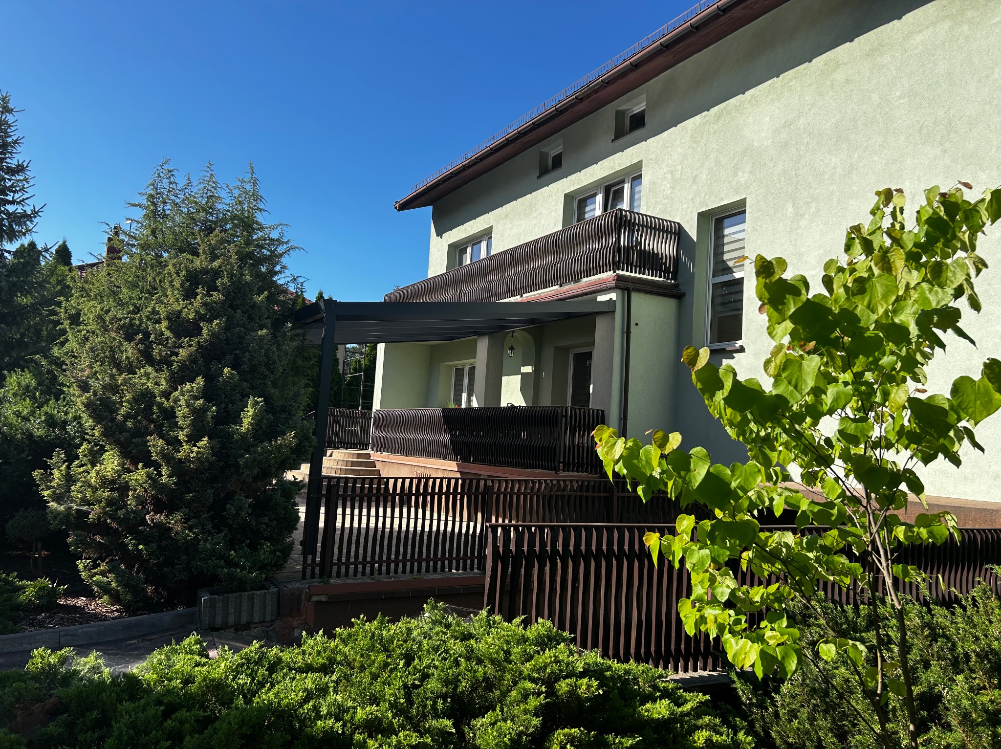 Dwukondygnacyjny dom z balkonem i tarasem z ciemną, nowoczesną pergolą. Widok z ogrodu w słoneczny dzień. Elementy drewniane i roślinność.