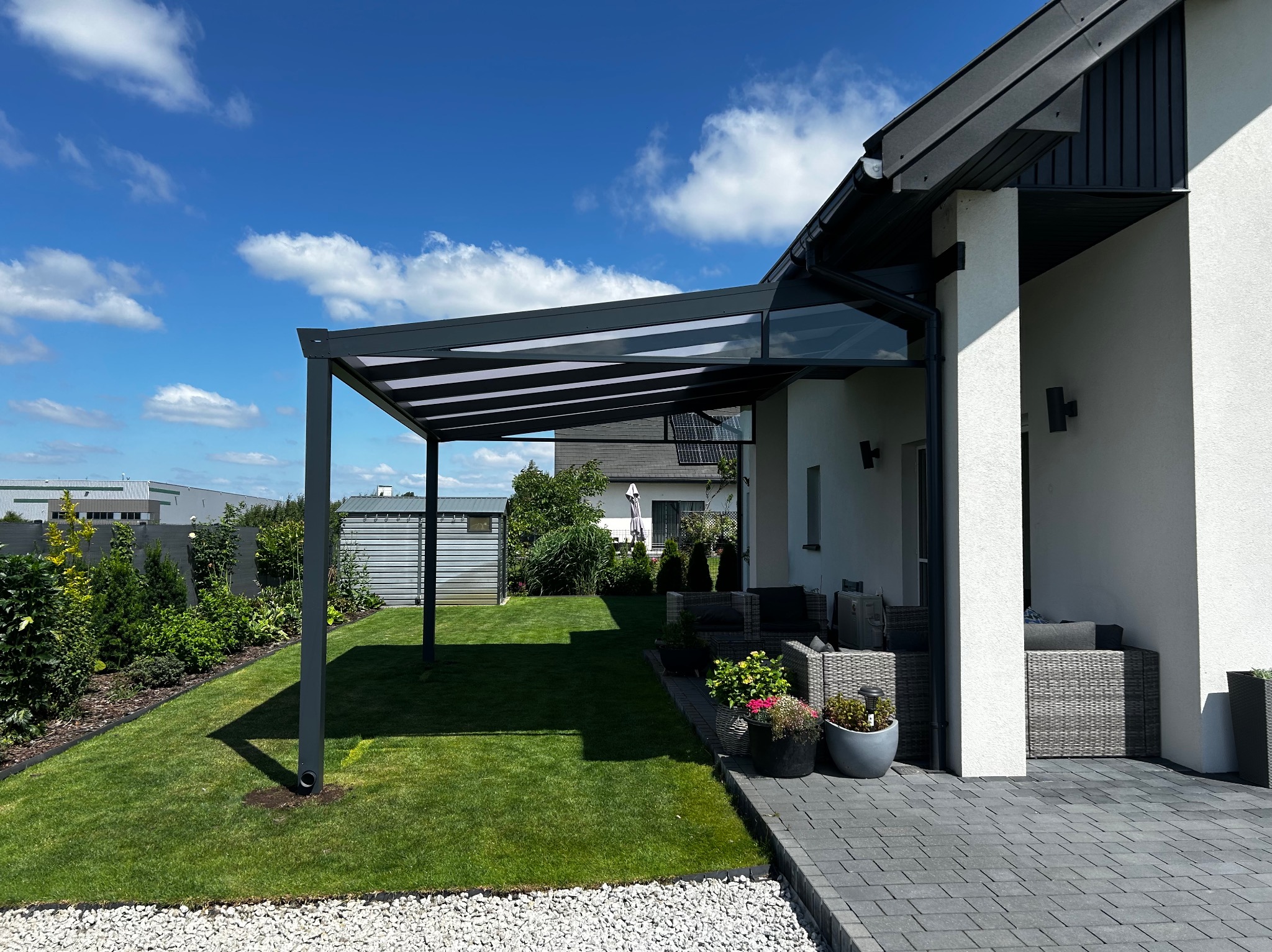 Nowoczesna pergola metalowa z zadaszeniem lamelowym, przylegająca do białego budynku. W tle zielony trawnik i błękitne niebo. Widoczne meble ogrodowe.