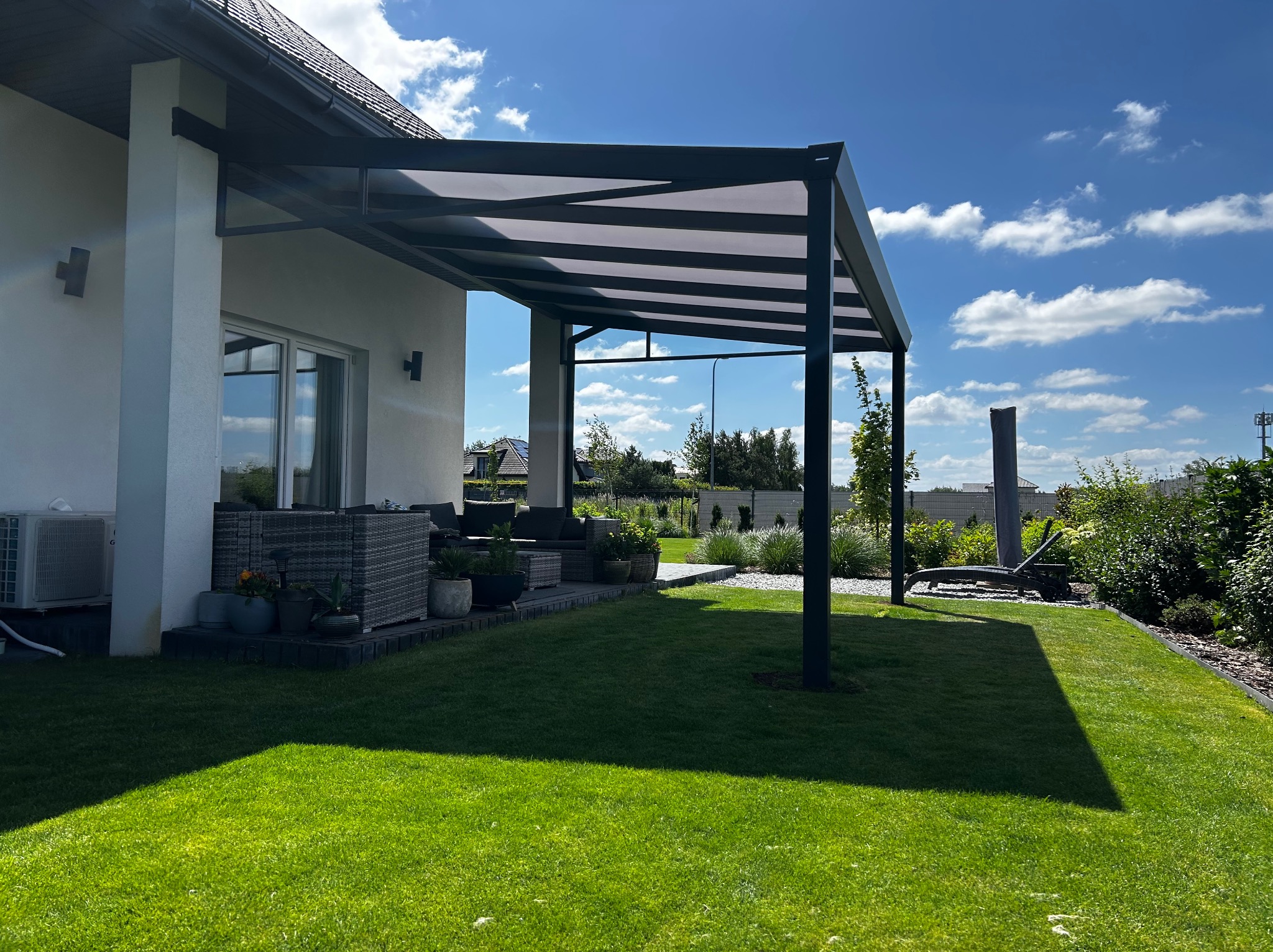 Nowoczesna pergola metalowa z zadaszeniem lamelowym przy białym domu, z widokiem na zadbany trawnik i meble ogrodowe w słoneczny dzień.