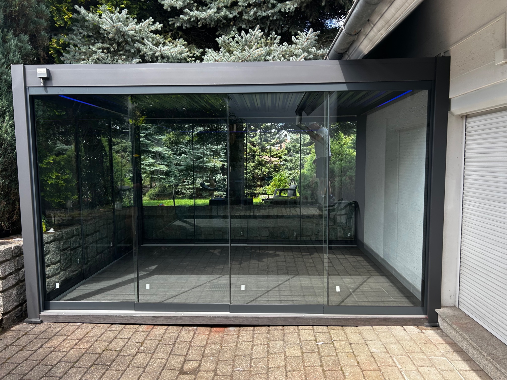 Szklana zabudowa tarasu z przesuwnymi panelami, widok na ogród. Nowoczesna konstrukcja z szarymi profilami, oświetlenie LED. Brukowana podłoga tarasu.