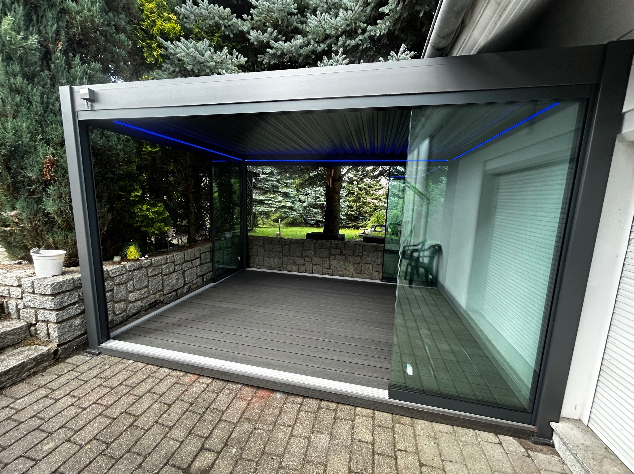 Szara, nowoczesna pergola z przeszkleniem i oświetleniem LED, widok z zewnątrz. Taras z desek kompozytowych, kamienne obrzeże i zieleń w tle.