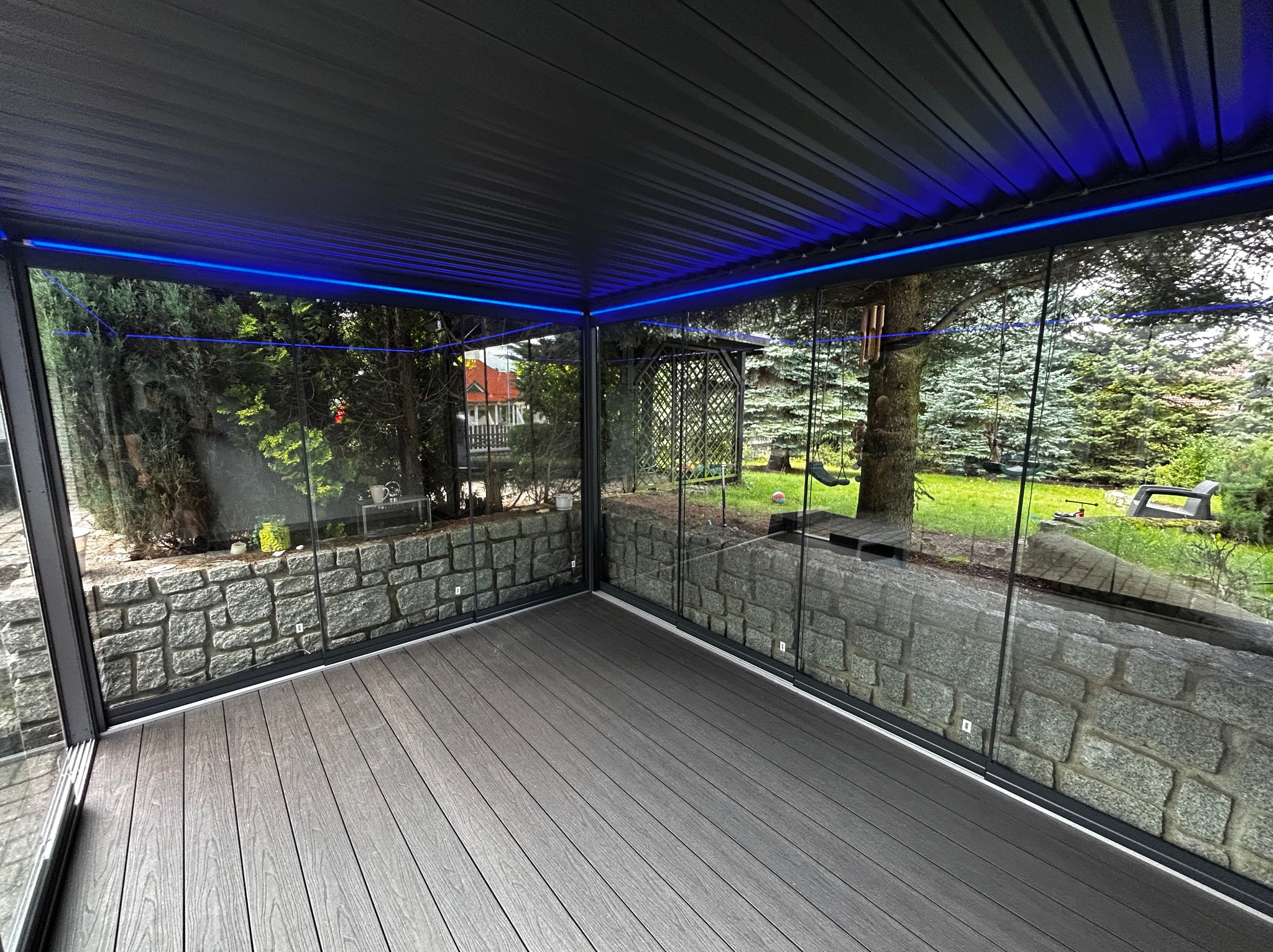 Nowoczesna zabudowa tarasu z szarych desek kompozytowych, przeszklonymi ścianami i podświetleniem LED w kolorze niebieskim. Widok na zielony ogród.