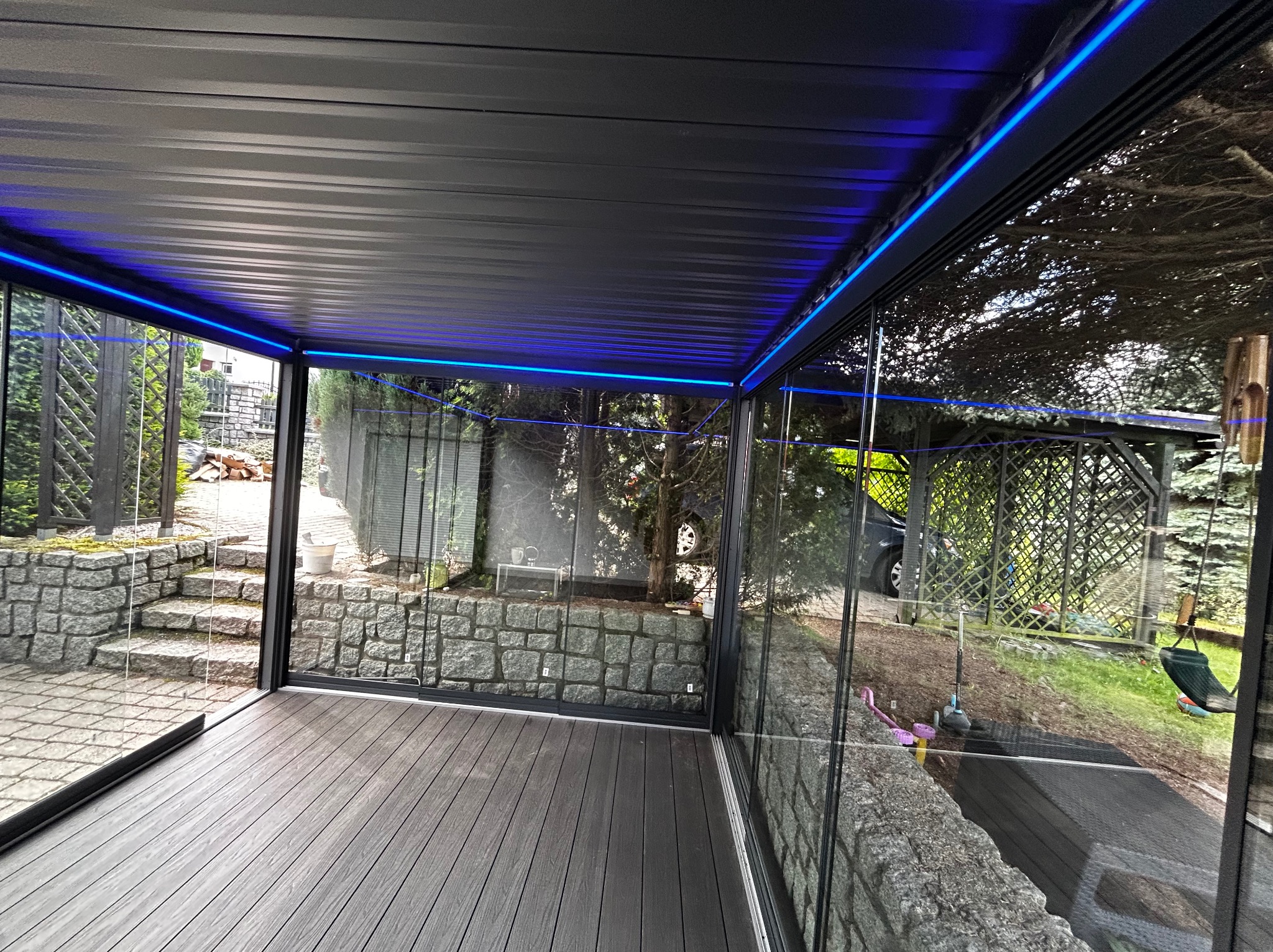 Nowoczesna zabudowa balkonu z szarym, kompozytowym tarasem i oświetleniem LED. Szklane ściany odbijają otoczenie, tworząc przestronny, osłonięty kącik.