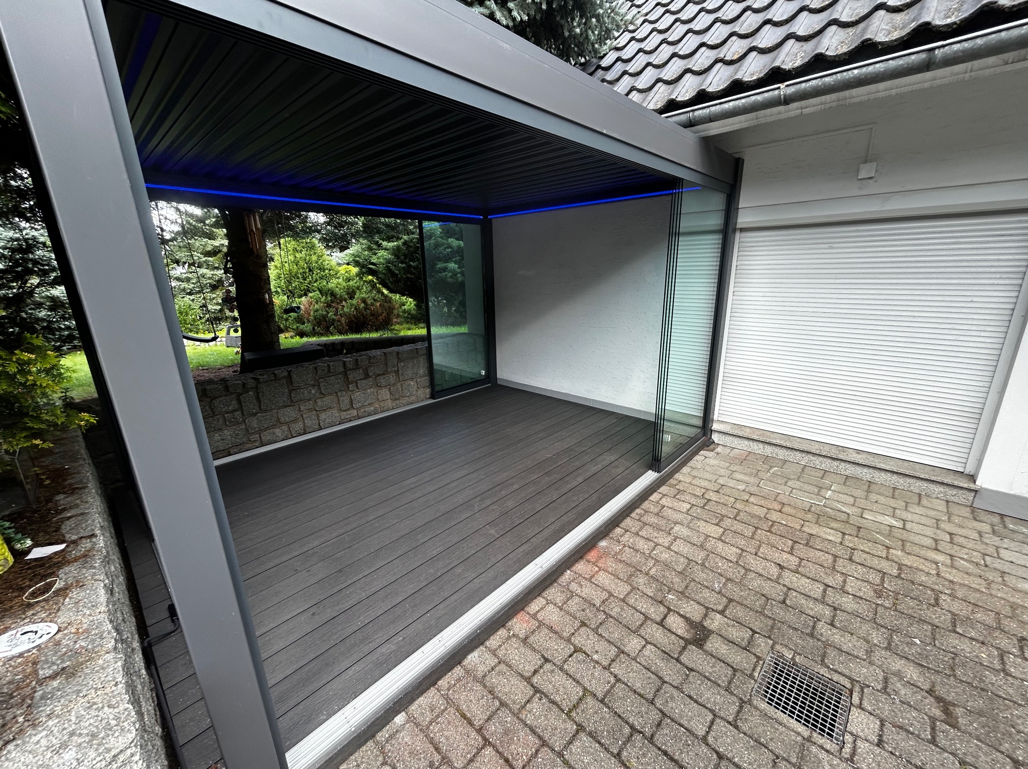 Nowoczesna, szara pergola z oświetleniem LED i szklanymi ścianami, przylegająca do budynku z roletą. Taras z desek kompozytowych.