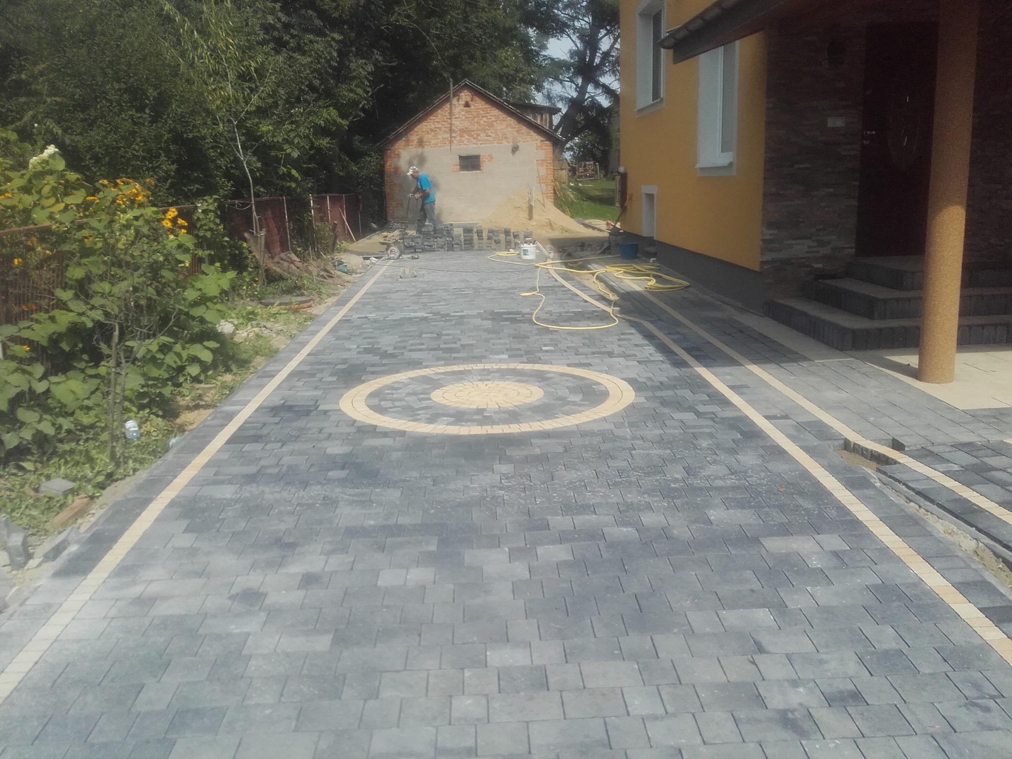 Wjazd do posesji wyłożony szarą kostką brukową z dekoracyjnym okręgiem w kolorze piaskowym; w tle pracownik układający kolejne kostki, widoczny budynek gospodarczy i fragment elewacji domu.