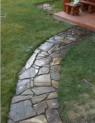 Ścieżka z nieregularnych kamieni w trawie, prowadząca do drewnianego tarasu z meblami ogrodowymi. W trawie widoczne dwa młotki.