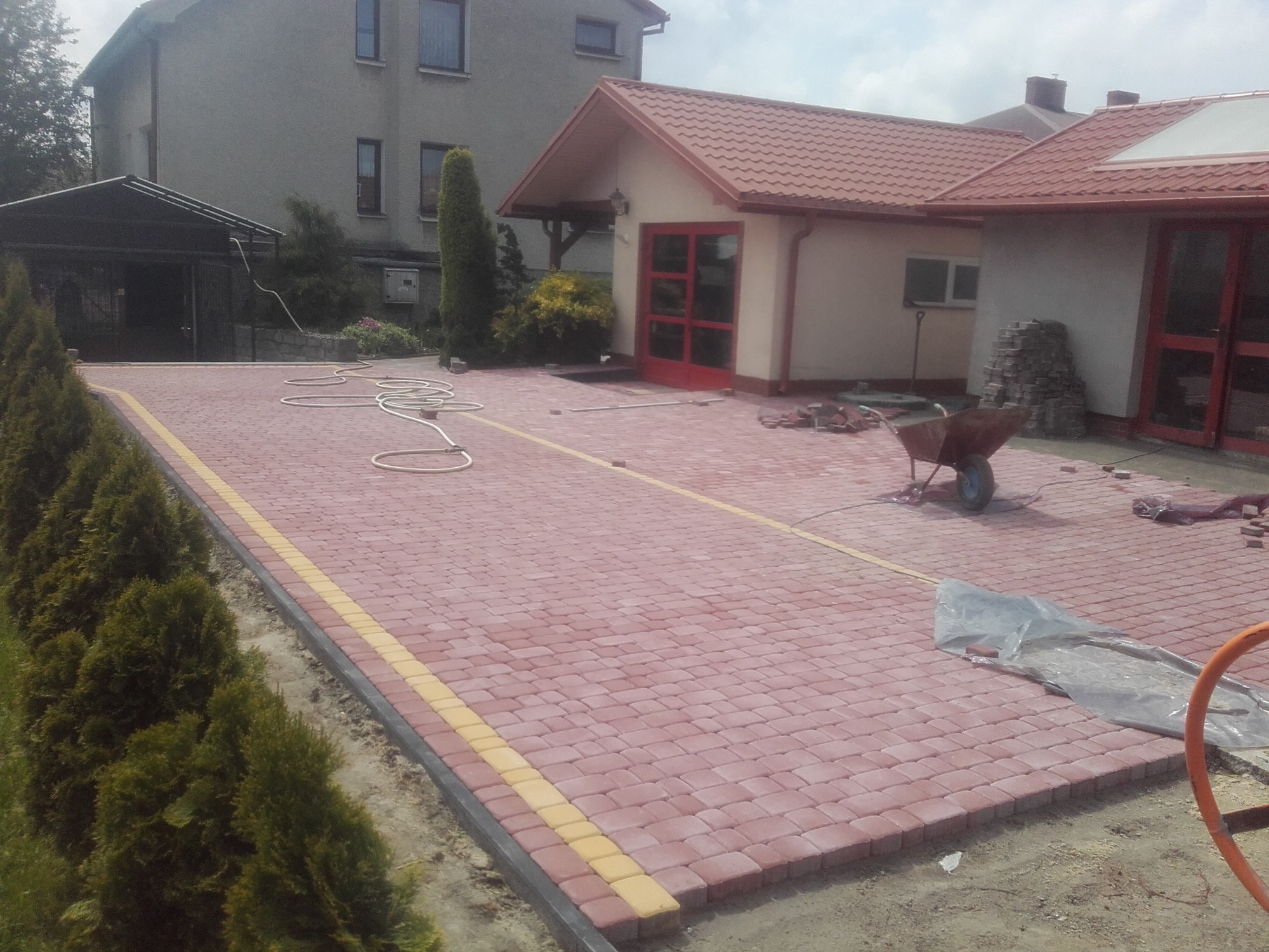 Czerwona kostka brukowa z żółtym obramowaniem, ciągnąca się wzdłuż rzędu zielonych drzew, w tle dom z czerwonym dachem, taczka i porozrzucane cegły wskazują na trwające prace.