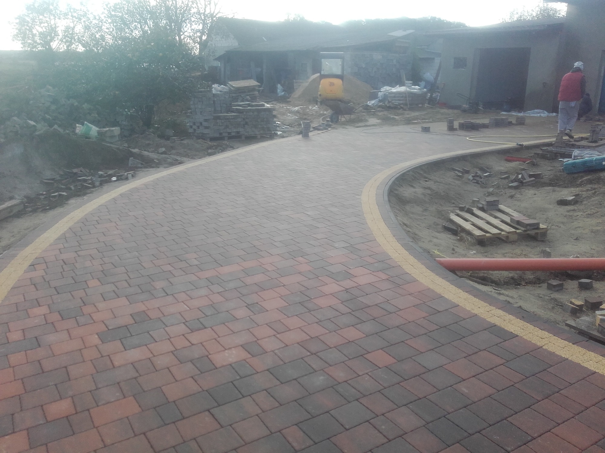 Wjazd z kostki brukowej w odcieniach brązu i szarości, z żółtym obramowaniem, w tle minikoparka i materiały budowlane.