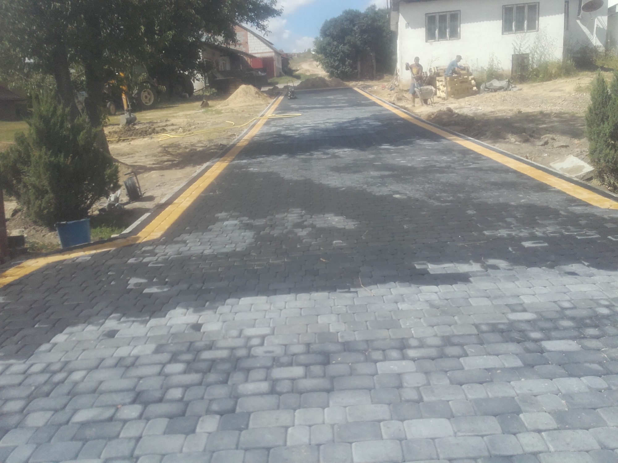 Ułożona ścieżka z szarej kostki granitowej z żółtymi obrzeżami, widoczni pracownicy i sprzęt budowlany w tle.