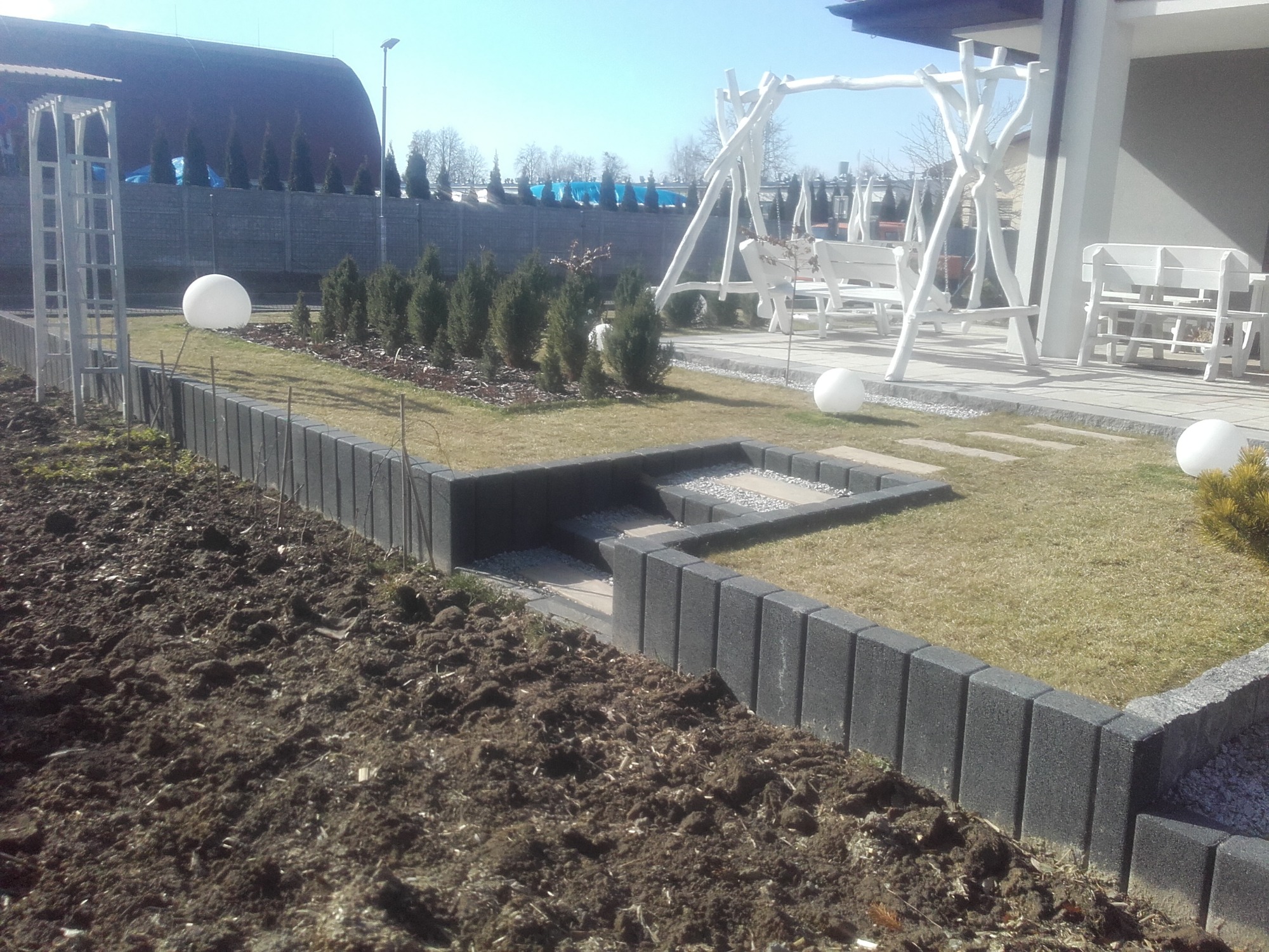 Fragment ogrodu w trakcie aranżacji: świeżo przekopana ziemia, obrzeża z betonowych bloczków, trawnik z nasadzeniami iglaków, białe kule dekoracyjne i biała, drewniana huśtawka w tle.