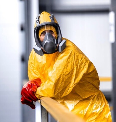 Osoba w żółtym kombinezonie ochronnym i masce, z czerwonymi rękawicami, oparta o metalową barierkę.