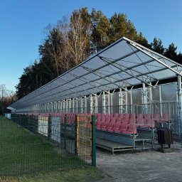 MR montaż Sp. z o.o. - Zadaszona trybuna z czerwonymi siedzeniami na boisku sportowym, widoczna przez metalowe ogrodzenie. Konstrukcja stalowa z przezroczystym zadaszeniem, drzewa w tle.