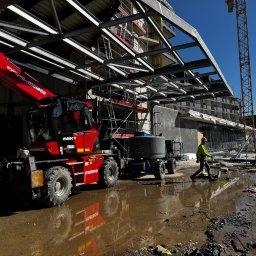 MR montaż Sp. z o.o. - Budowa hali w toku: stalowa konstrukcja, żuraw i podnośniki na mokrym placu budowy. Pracownik w żółtej kamizelce przechodzi obok maszyn.