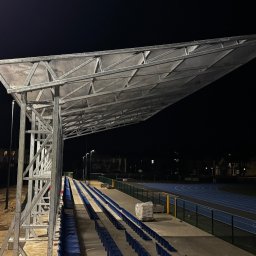 MR montaż Sp. z o.o. - Stalowa konstrukcja zadaszenia trybun na stadionie sportowym nocą. Widoczne niebieskie siedzenia i bieżnia. Oświetlenie uliczne w tle.