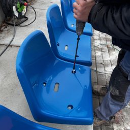 MR montaż Sp. z o.o. - Montaż niebieskich krzesełek stadionowych. Widoczny pracownik w trakcie przykręcania siedziska w rzędzie, używając wkrętarki. W tle zwinięty kabel zasilający.
